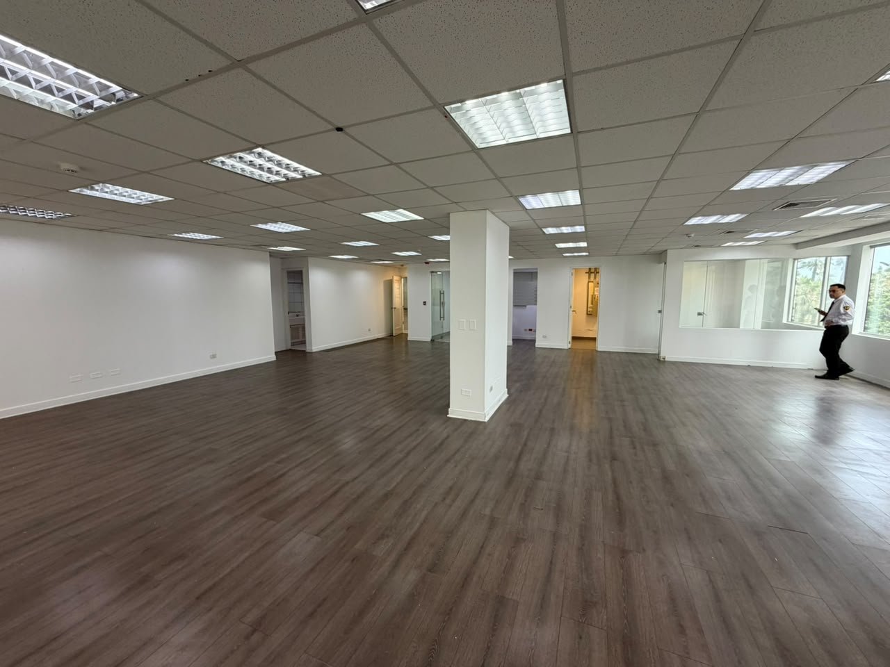 Empty commercial office space with dark wood flooring, white columns, fluorescent ceiling lights, and a person visible in the background