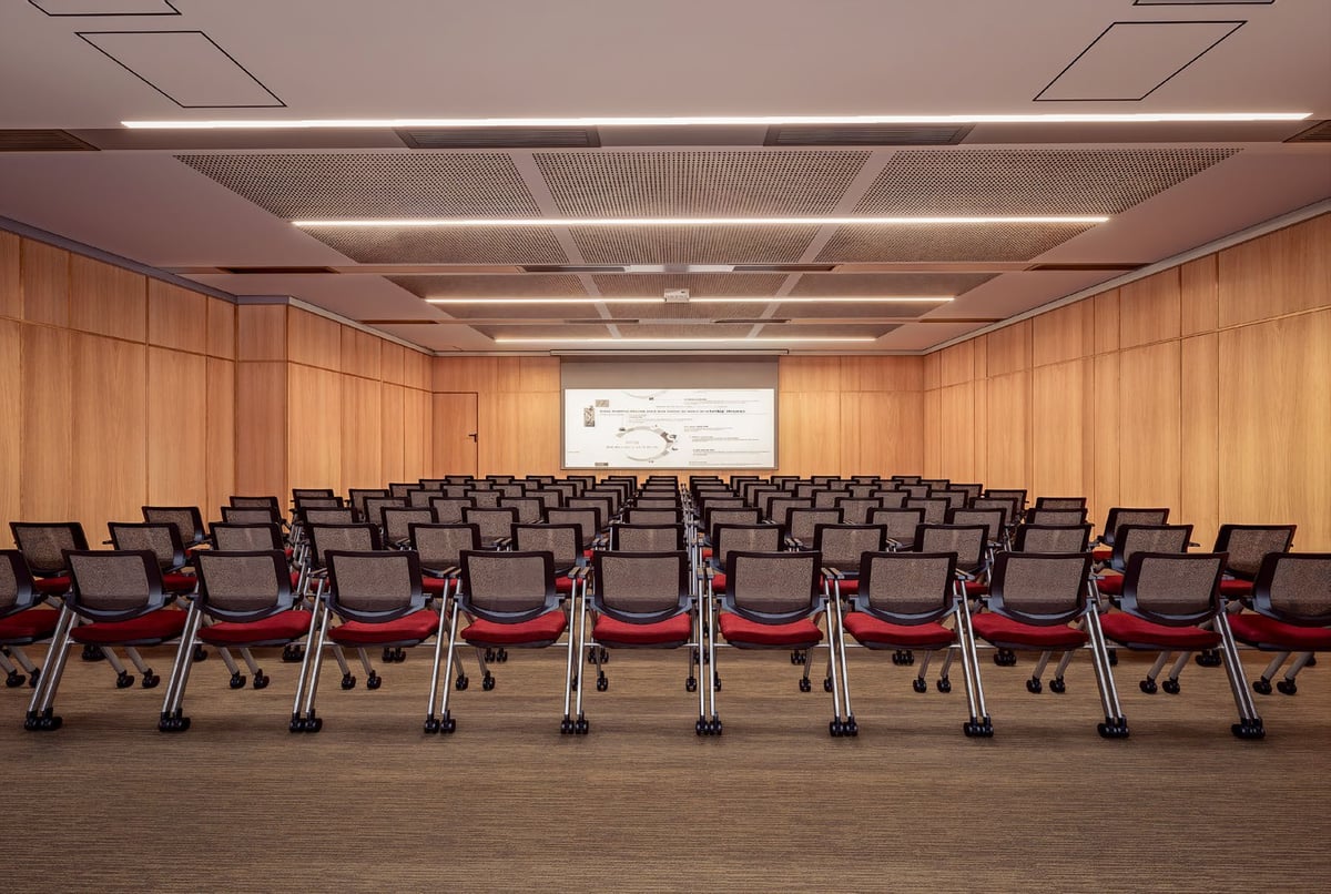 Empty modern conference room with rows of red and black mesh chairs facing a projection screen on wood-paneled wall