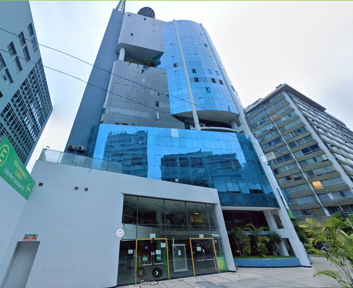 Modern multi-story building with blue glass panels and white lower facade, ground-level storefront entrance, surrounded by other residential buildings