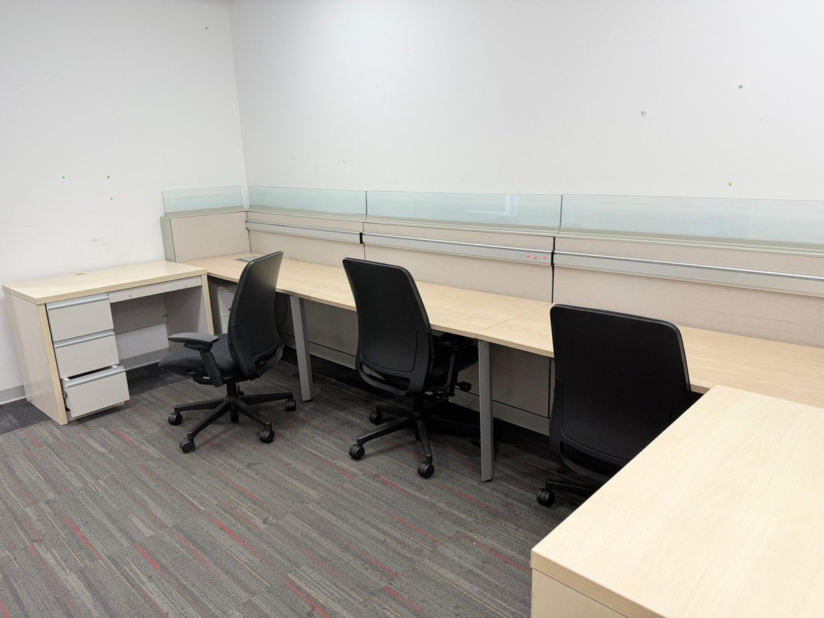 Modern office workstation with light wood desks, black office chairs, glass dividers, and gray carpeting