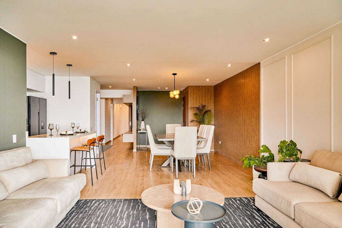 Modern open-concept apartment with cream sofas, dining area, white kitchen, wooden accents, and potted plants throughout