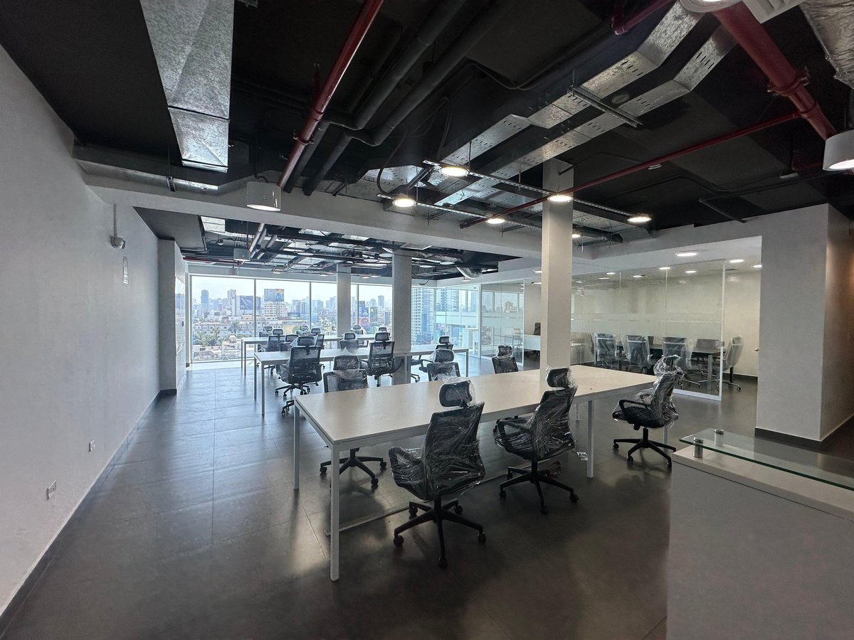 Modern open office space with mesh chairs at a white table, industrial ceiling with exposed ducts, and floor-to-ceiling windows overlooking a city skyline