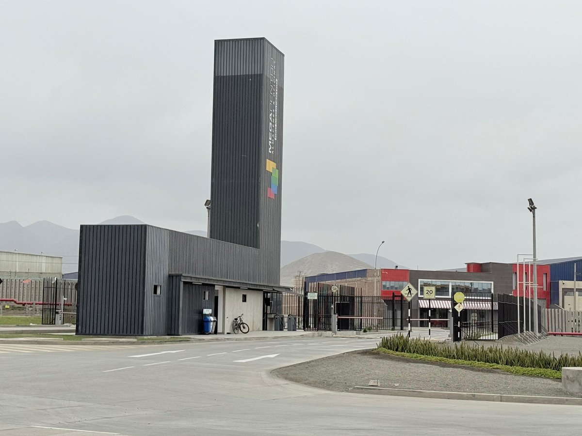 Modern industrial complex with tall vertical striped metal tower building, lower adjacent structure, and surrounding commercial facilities in a paved courtyard area with mountains in the background.