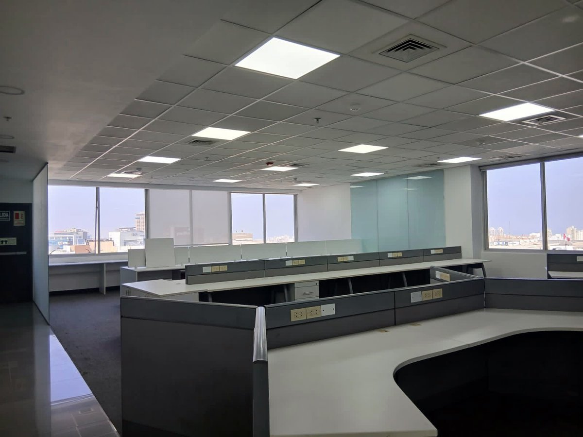 Empty modern office space with black desks, large windows, fluorescent ceiling lights, and cityscape view