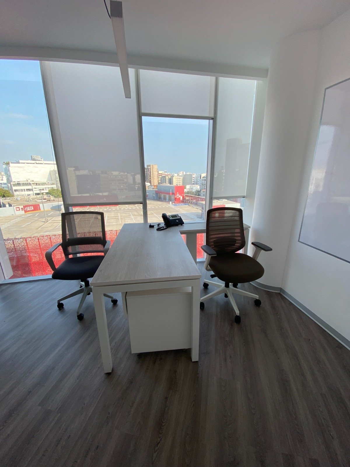 Modern office space with two black mesh chairs at a white desk, large windows overlooking a city skyline, and hardwood flooring.