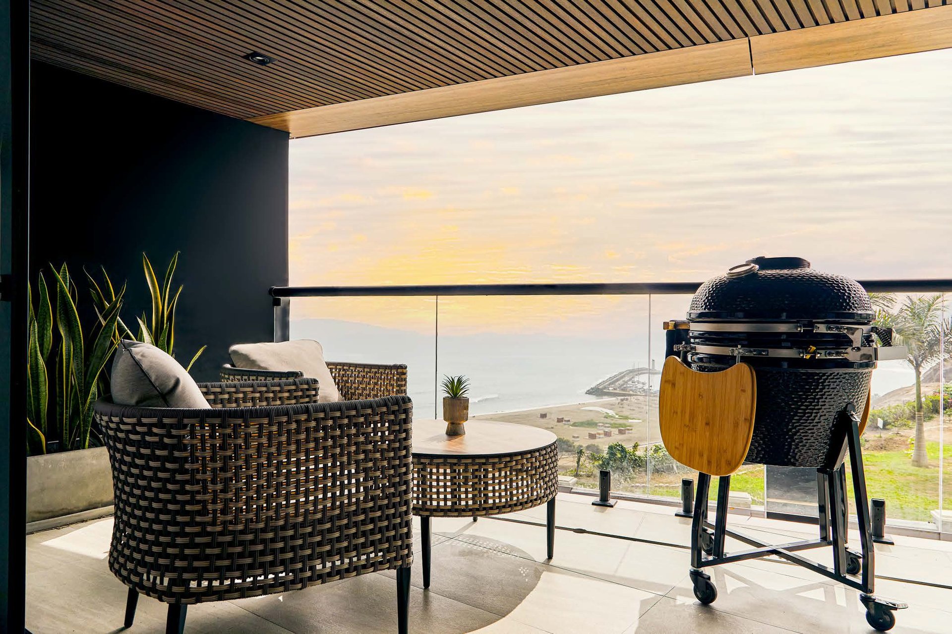 Modern outdoor patio with black woven armchair, round side table, and black dome grill overlooking scenic hillside landscape