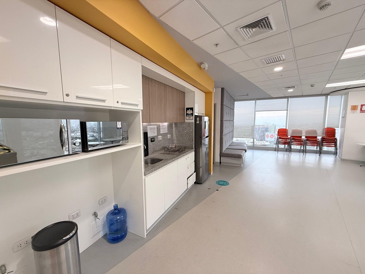 Modern medical office break room with white cabinetry, yellow accent beam, kitchenette, trash bin, water cooler, and red chairs visible in waiting area