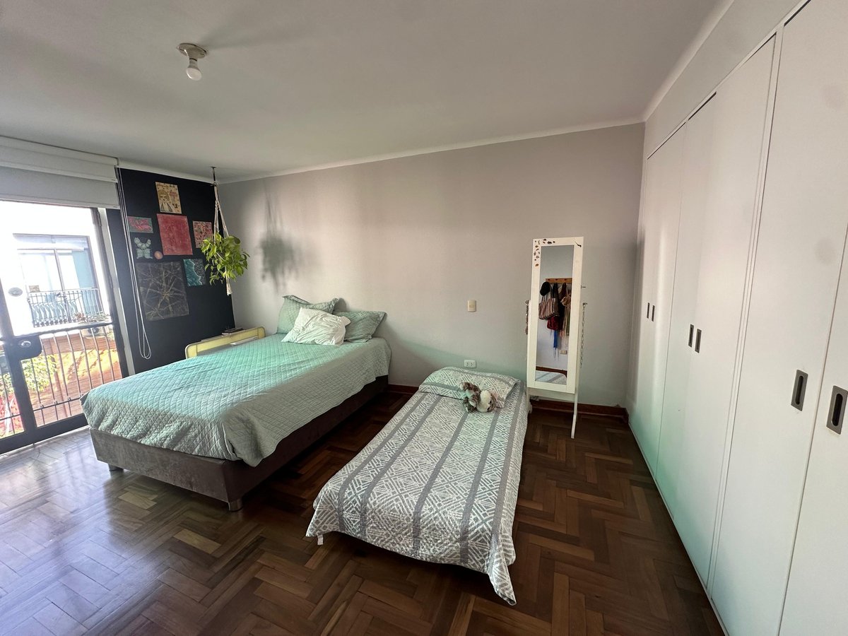 A bright bedroom with two beds, white wardrobes, and wooden herringbone flooring, with a view of a balcony through the window.
