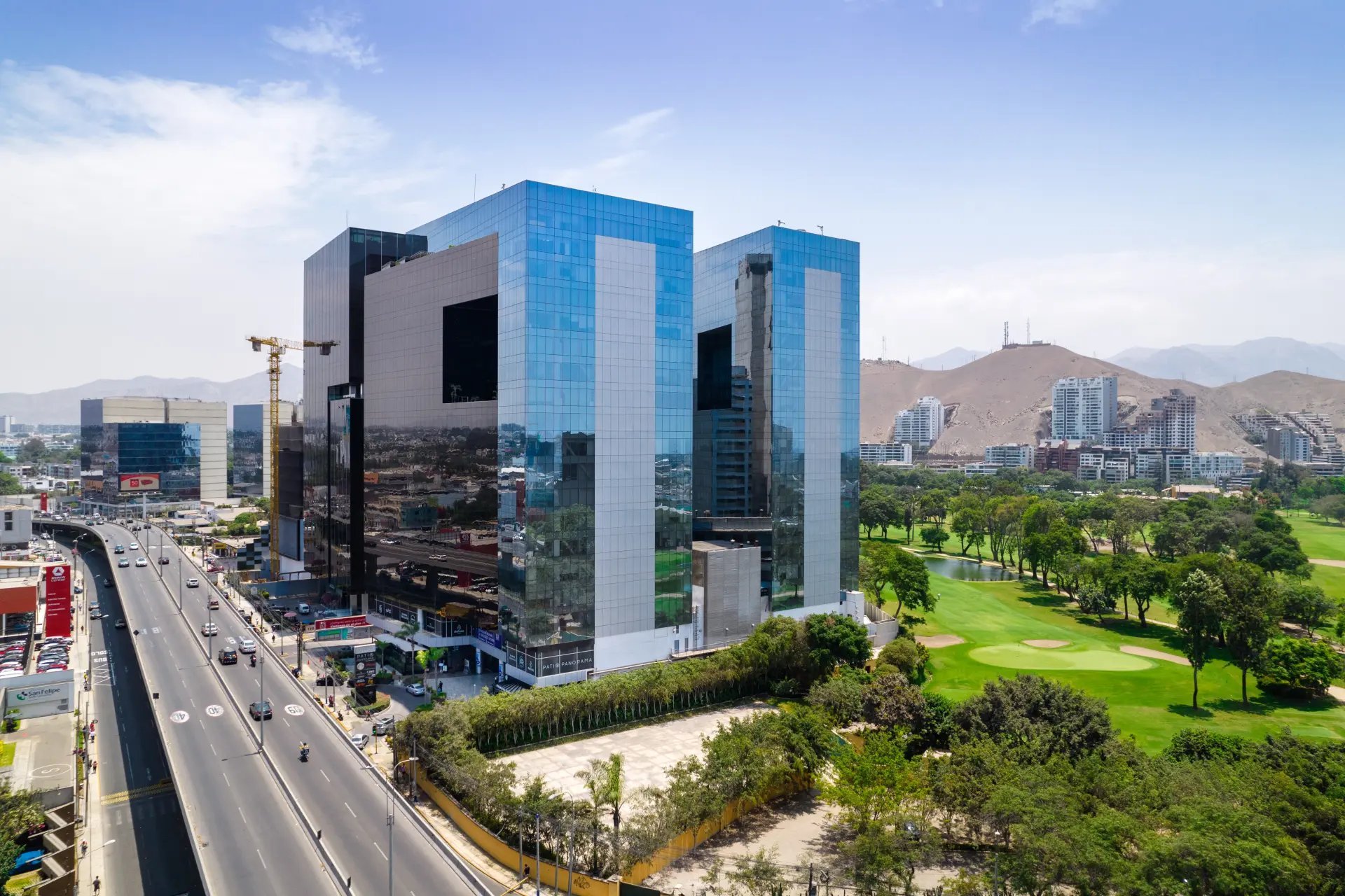 Modern glass office building complex with blue-tinted facades overlooking a green park and distant city skyline with mountains