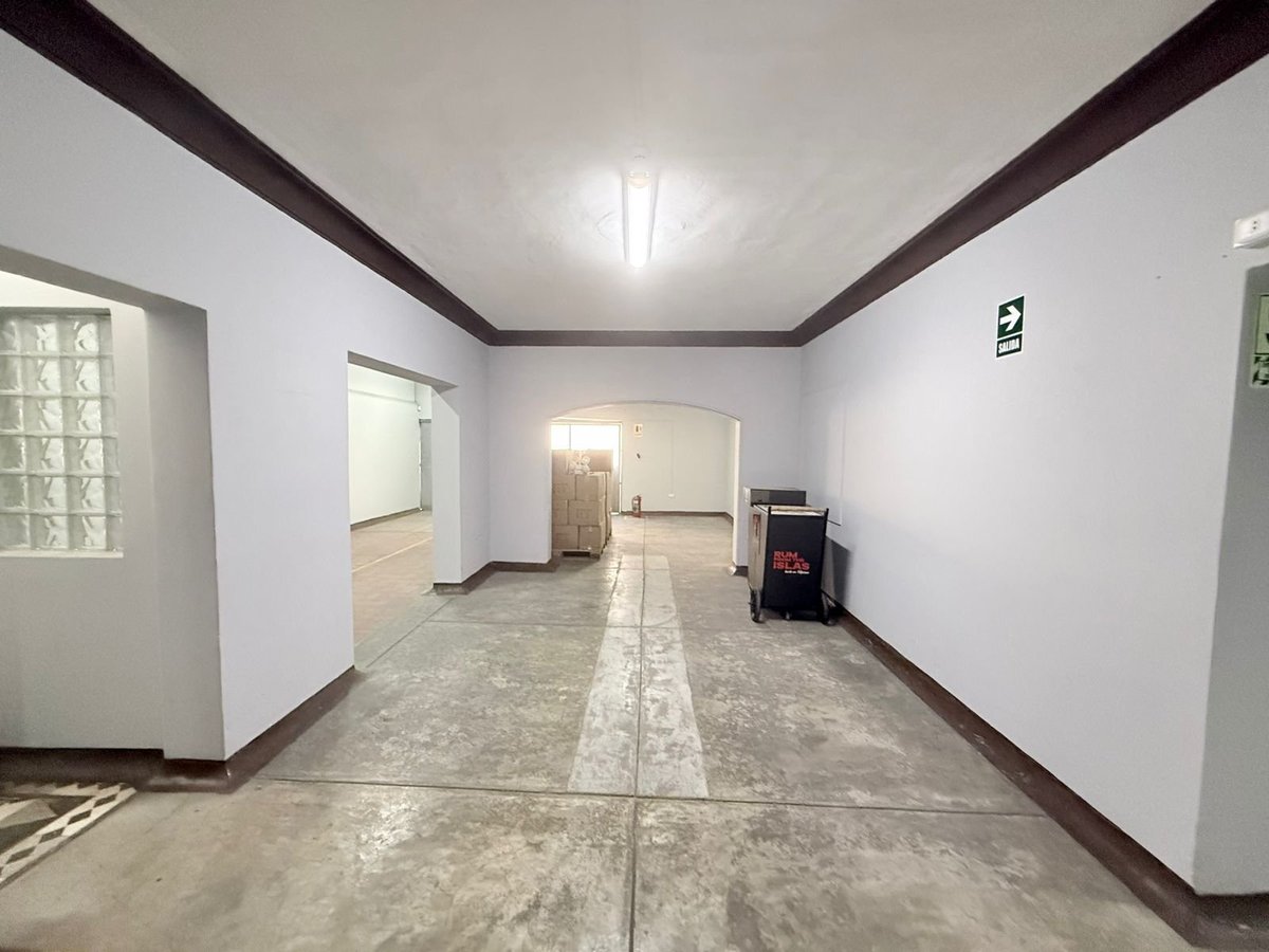 Empty interior room with white walls, dark wooden ceiling beams, concrete floor, and doorways leading to adjoining spaces.