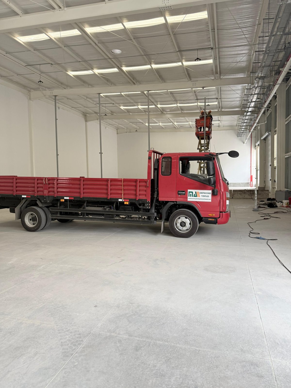 Red truck with cargo bed parked in a modern industrial warehouse with exposed beam ceiling and bright lighting.