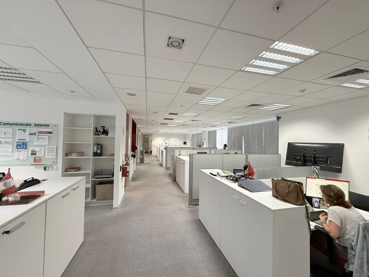 Modern office space with white cubicles, fluorescent ceiling lights, and clean gray flooring stretching toward the back of the room.