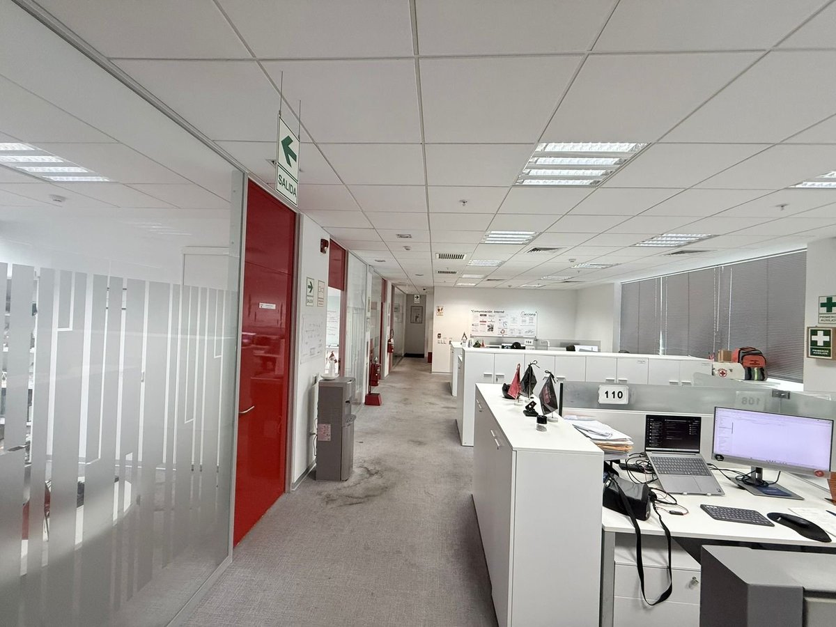Modern office interior with white drop ceiling, red emergency doors, white cubicles, computer workstations, and fluorescent lighting