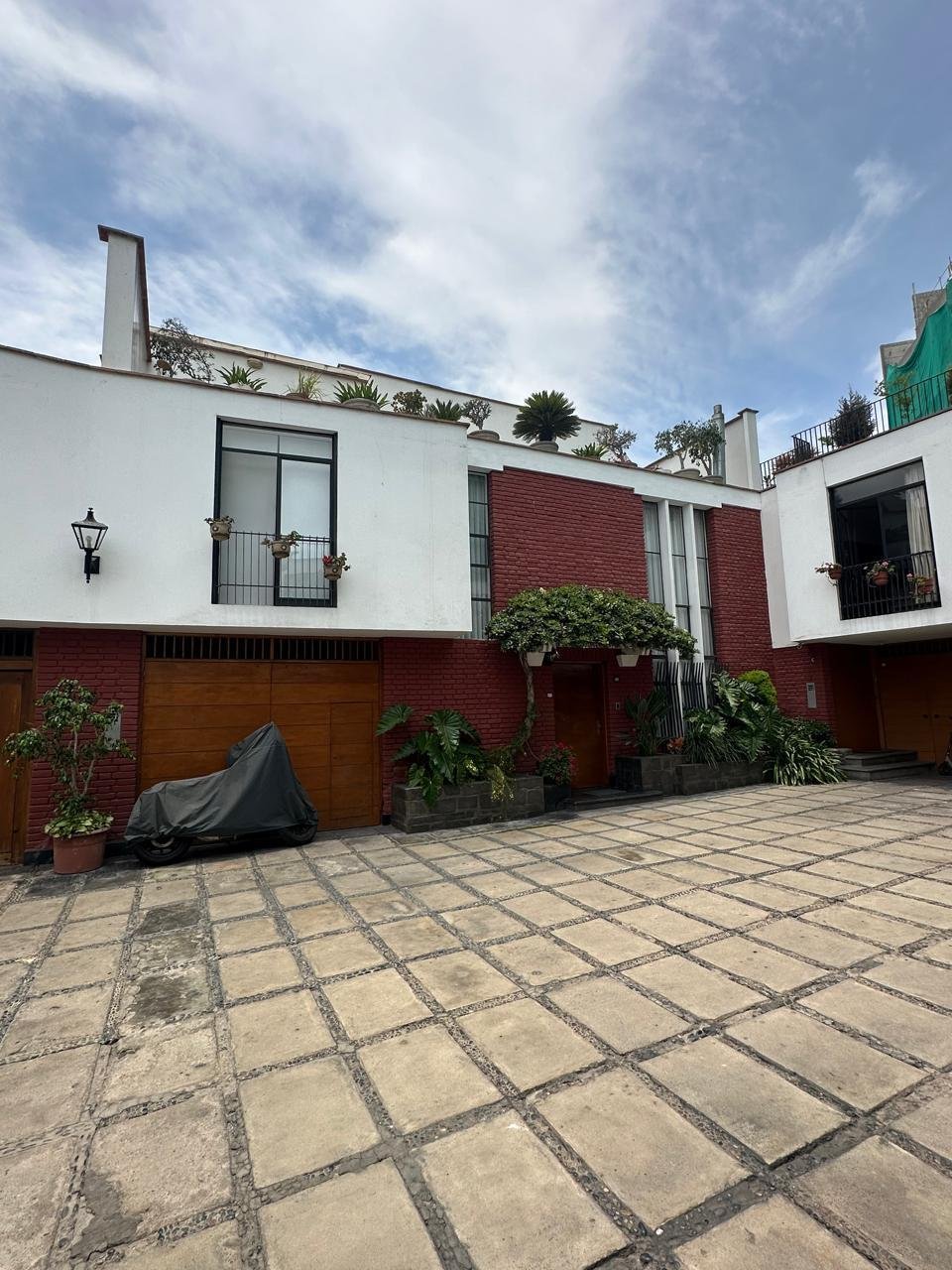 Modern white and red brick residential building with courtyard patio, wooden garage doors, and climbing plants under blue sky