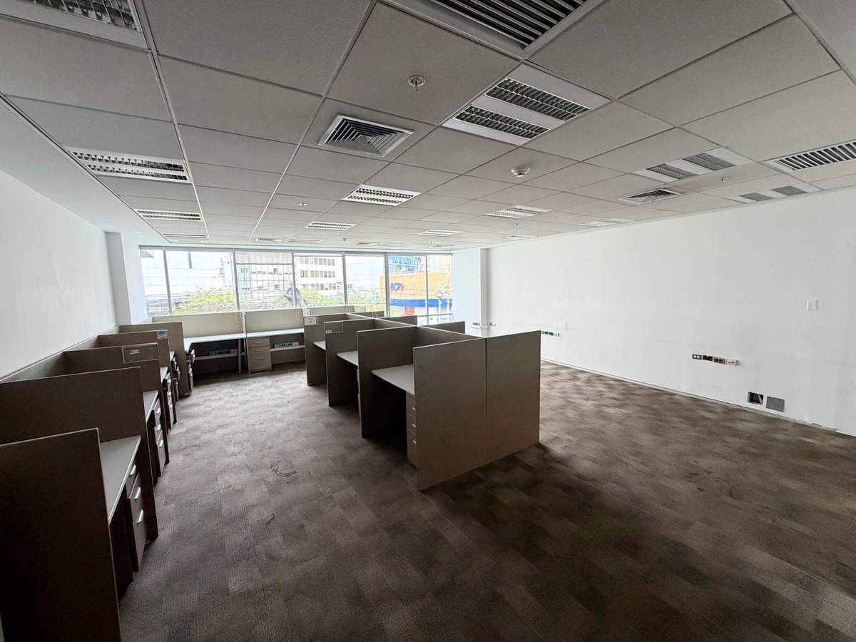 Empty modern office space with brown desks, drop ceiling, fluorescent lighting, and large windows overlooking a cityscape