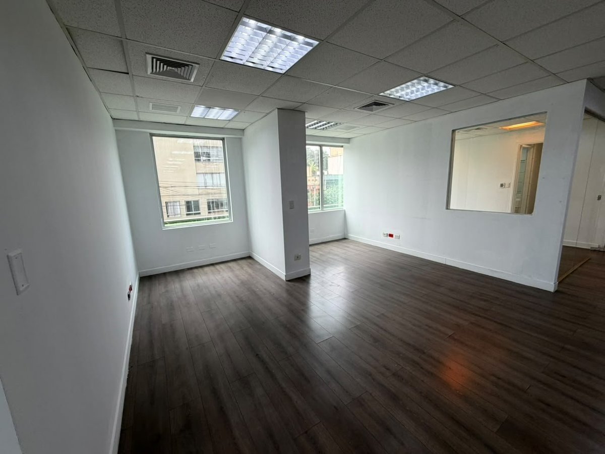 Empty office space with dark wood floors, white walls, fluorescent ceiling lights, and windows overlooking a city street.