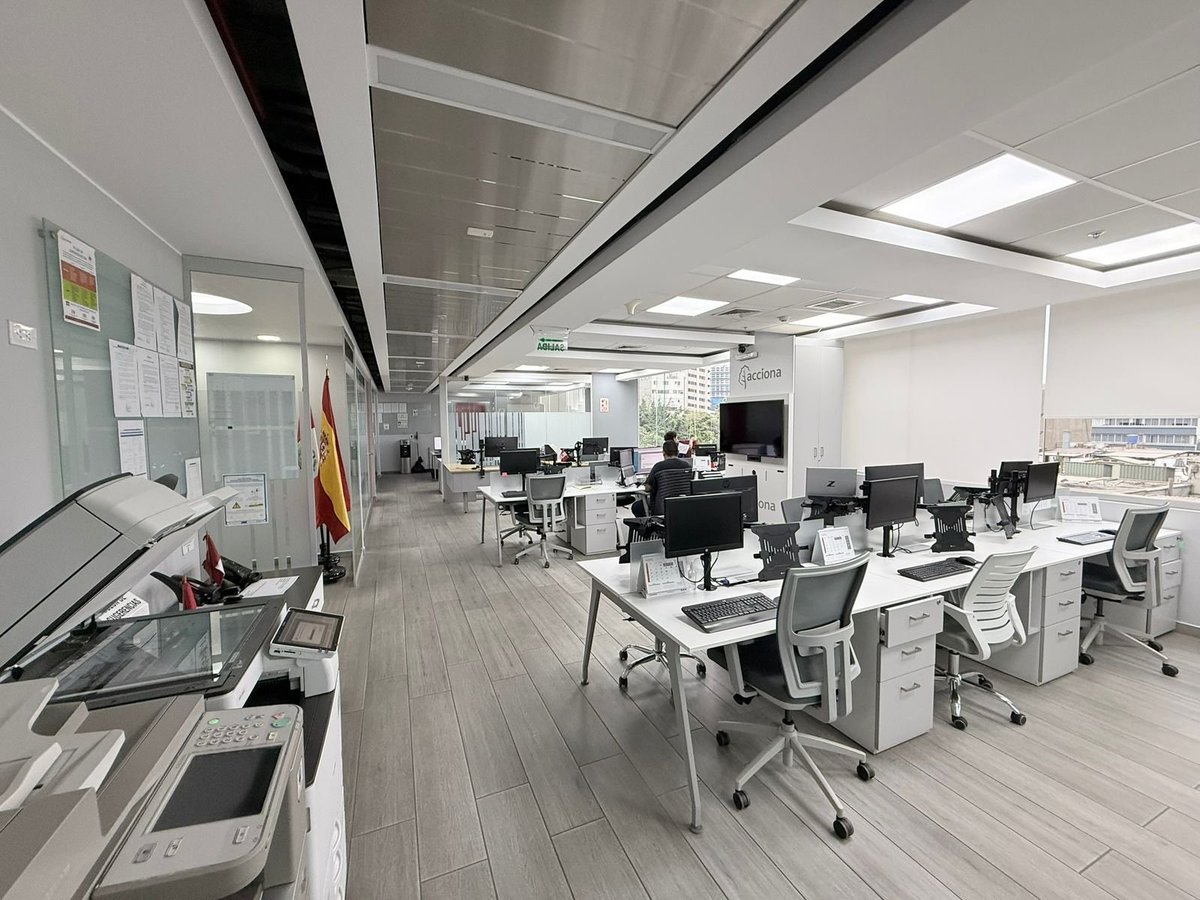 Modern open-plan office with white desks, ergonomic chairs, and computer monitors under bright ceiling lights and skylights