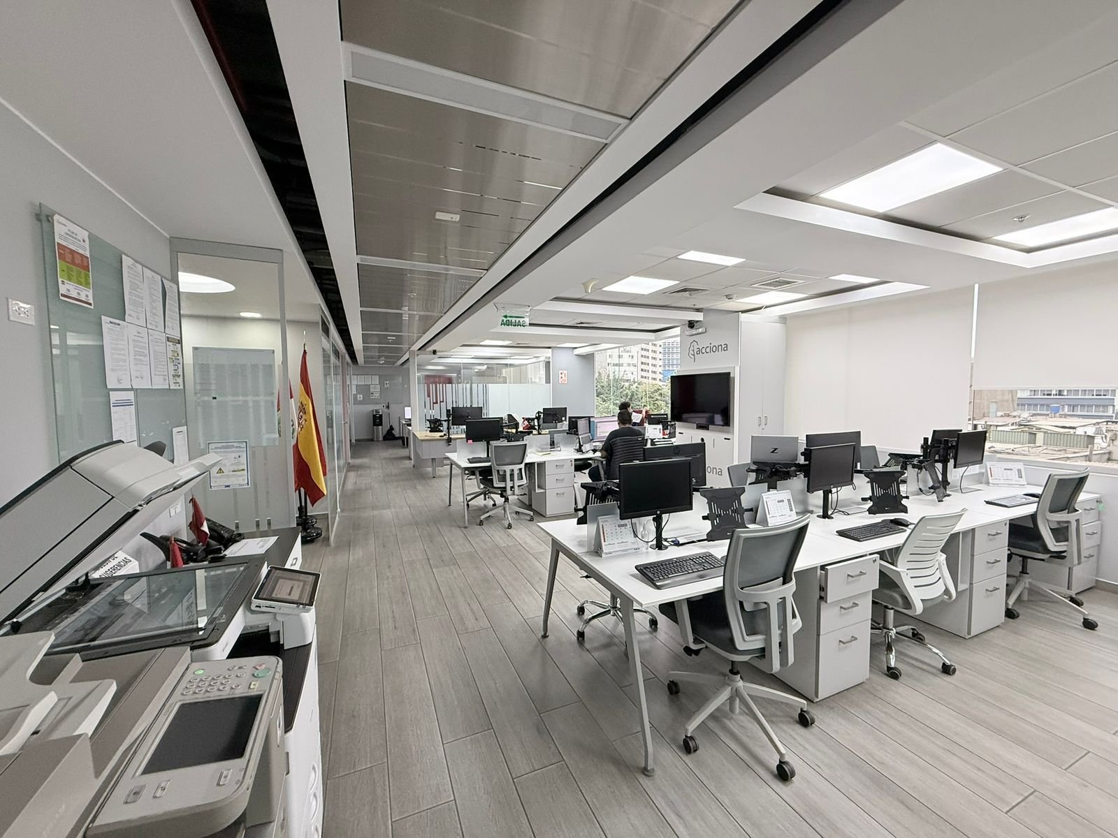 Modern open-plan office with white desks, ergonomic chairs, and computer monitors under bright ceiling lights and skylights