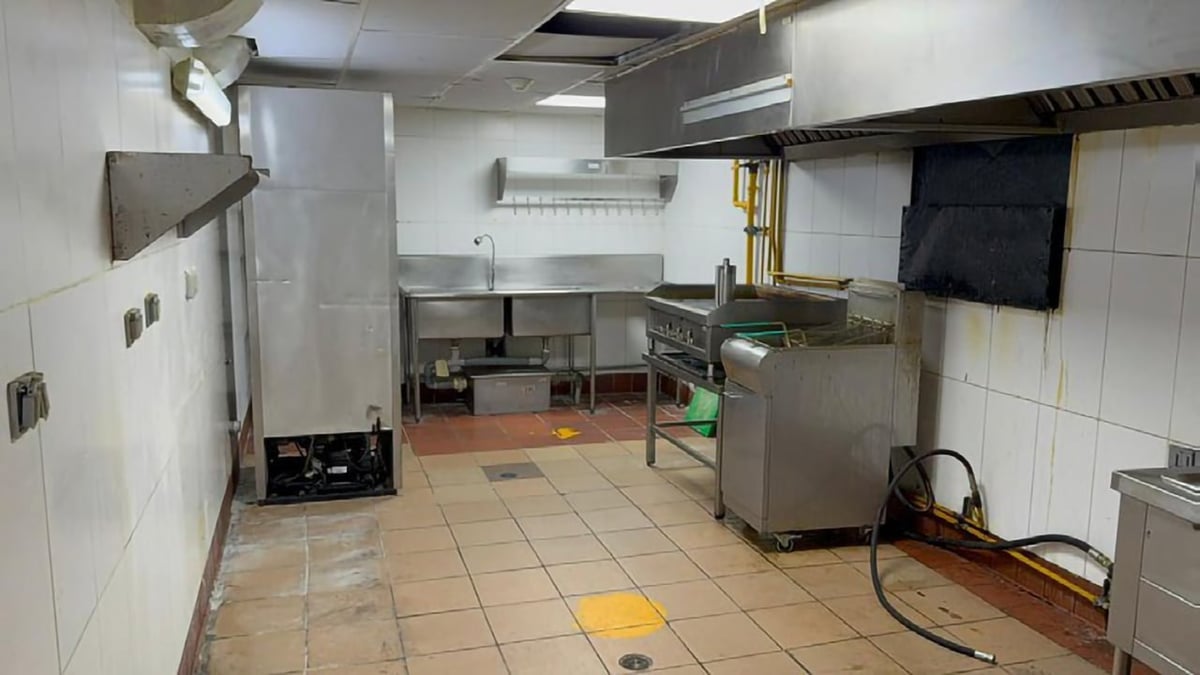 Empty commercial kitchen with stainless steel equipment, white tiled walls, and terracotta tiled floor