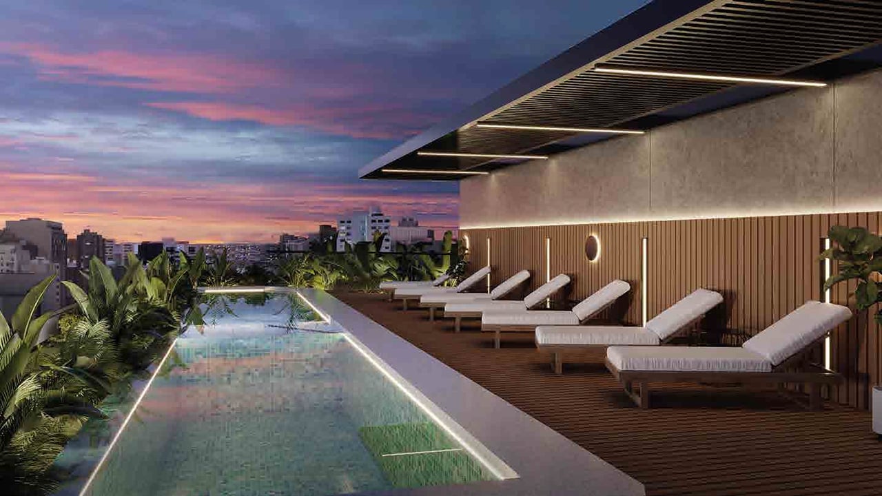 Modern rooftop lounge at sunset with illuminated pool, loungers, and city skyline in the background