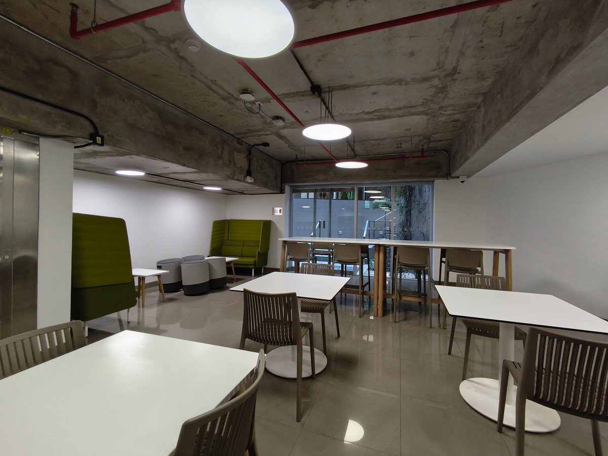Modern office break room with white tables, wooden chairs, olive green privacy screens, and concrete ceiling with skylights