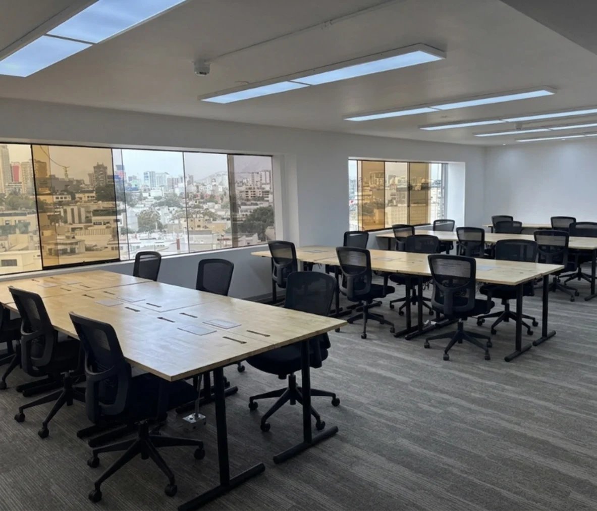 Modern open office space with white tables, black ergonomic chairs, and floor-to-ceiling windows overlooking a city skyline