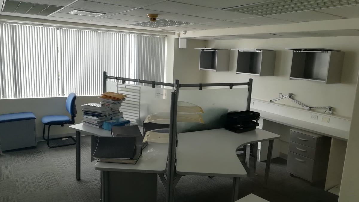 Modern office break room with a bed, desk, chair, and shelving. Vertical blinds on windows provide natural light