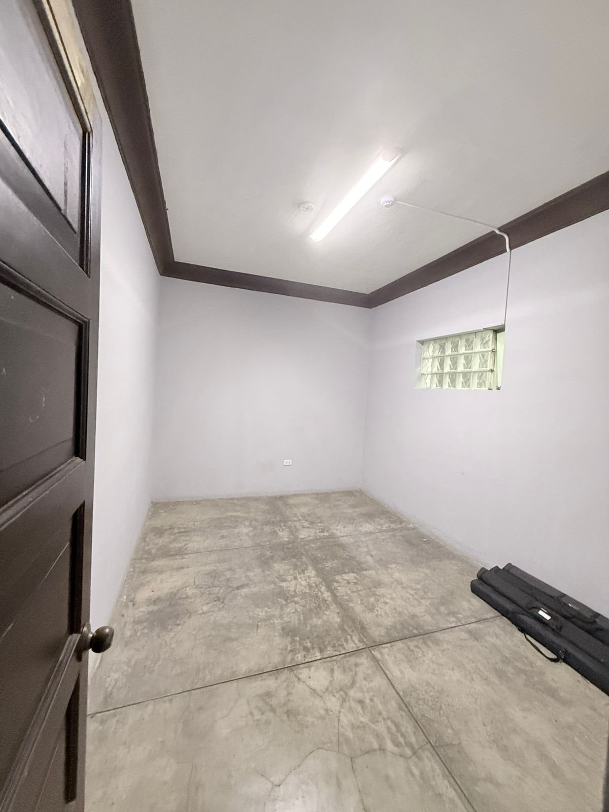 Empty room with white walls, dark wooden trim, concrete floor, fluorescent ceiling light, and a small high window