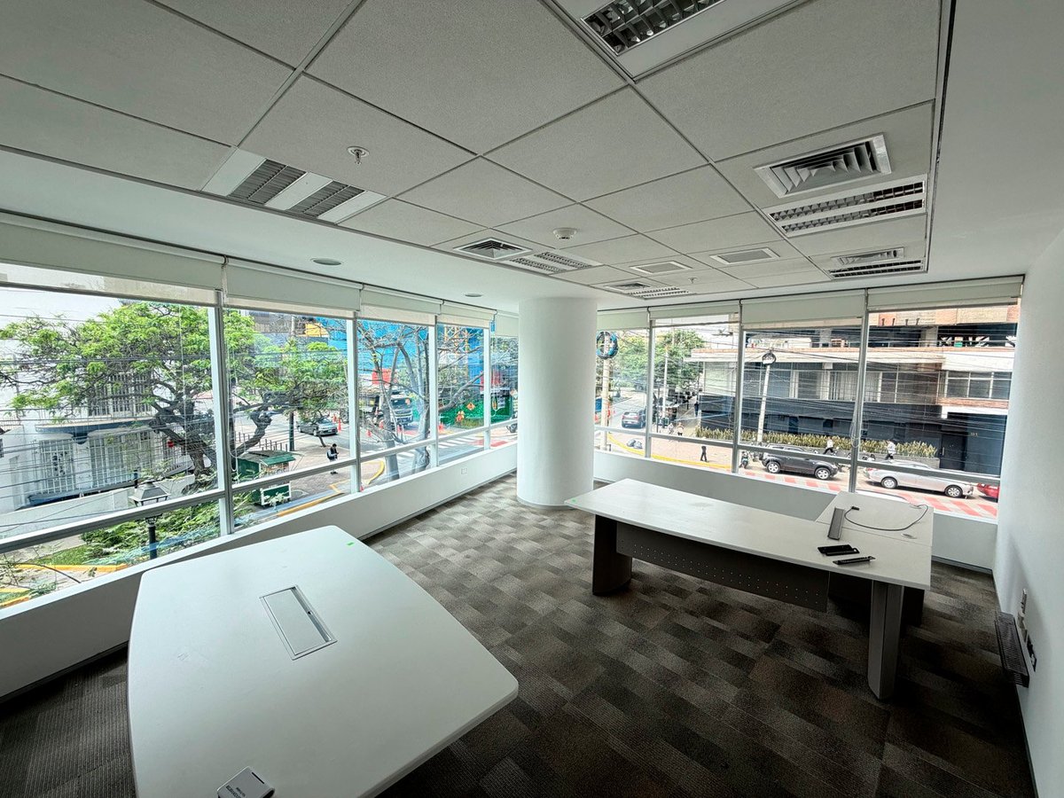 Empty modern office space with large windows overlooking city streets, white desk, and dropped ceiling with fluorescent lighting
