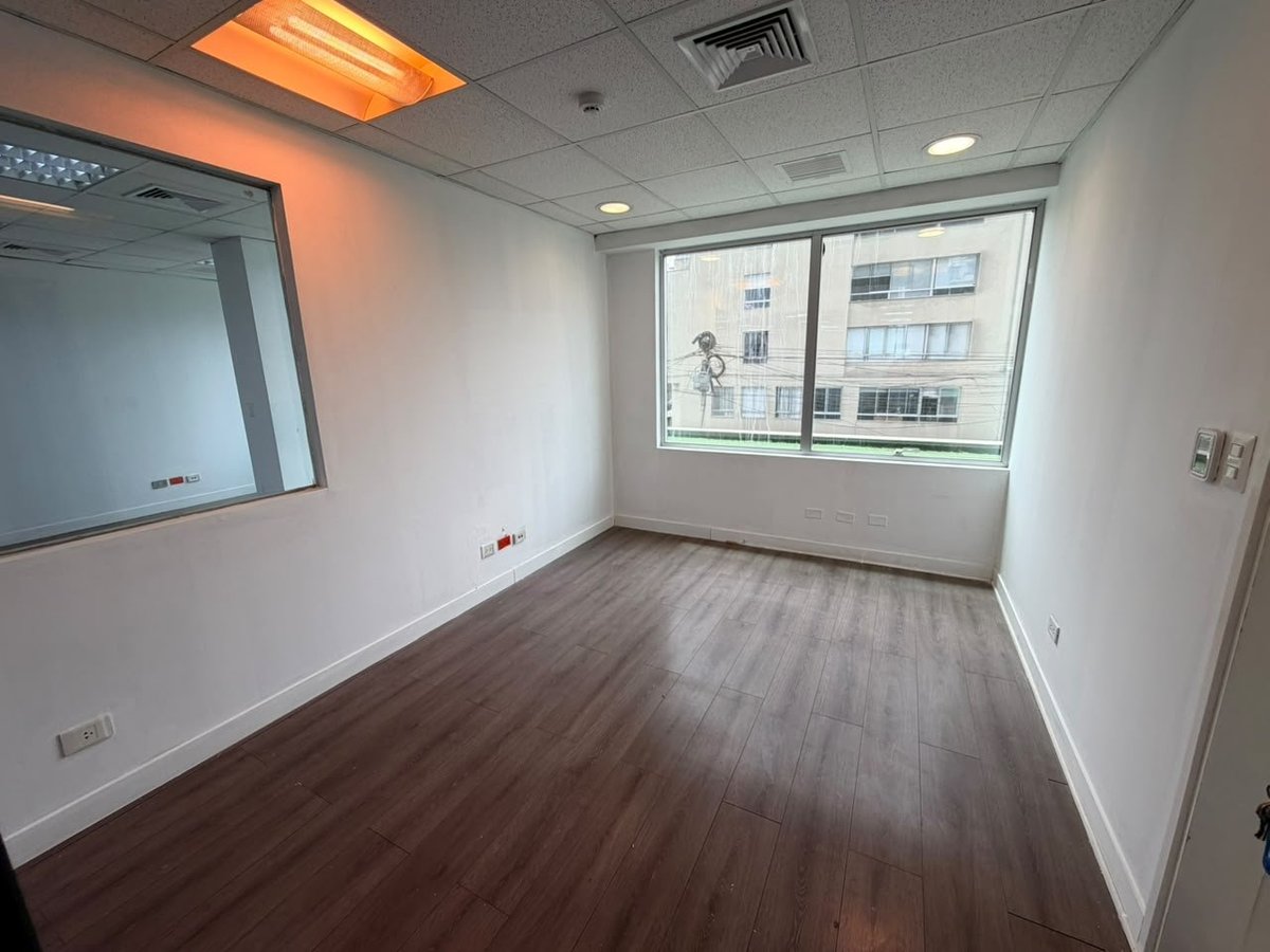 Empty office or commercial space with dark wood flooring, white walls, large window overlooking city buildings, and ceiling lights.
