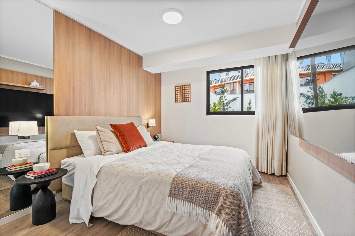 Modern bedroom with wooden feature wall, white bedding with red accent pillow, nightstands, and large windows with curtains.