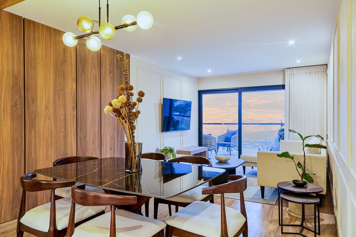 Modern luxury apartment interior with wooden dining area, blue upholstered seating, floor-to-ceiling windows overlooking a sunset cityscape
