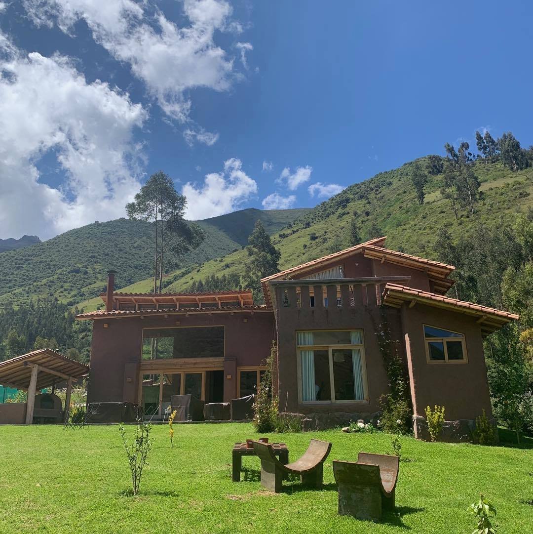 Modern brown wooden house with large windows on a green lawn, with picnic table and mountains in the background under blue sky