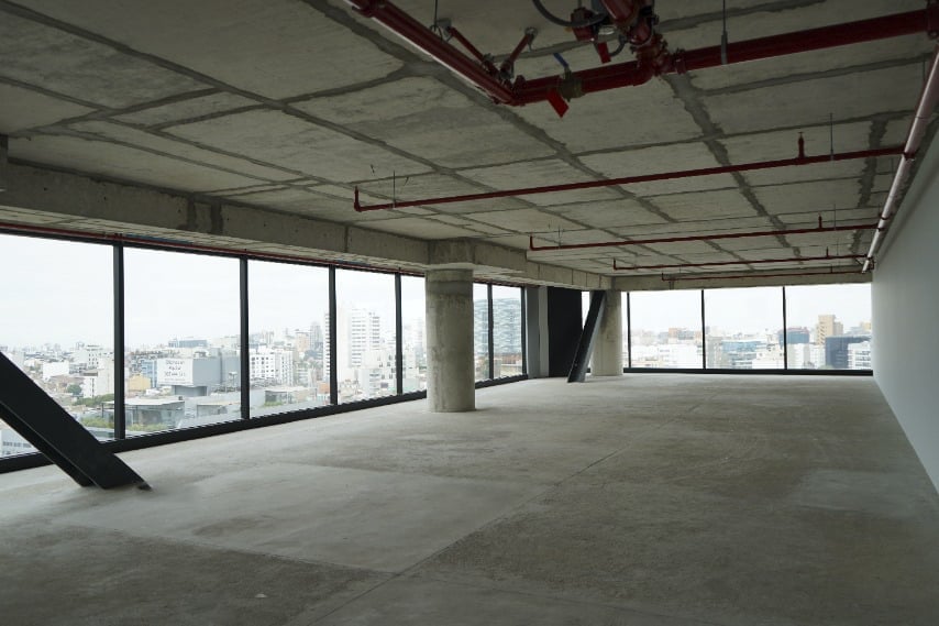 Empty modern office or commercial interior space with large windows overlooking city skyline, concrete floors, and exposed ceiling infrastructure