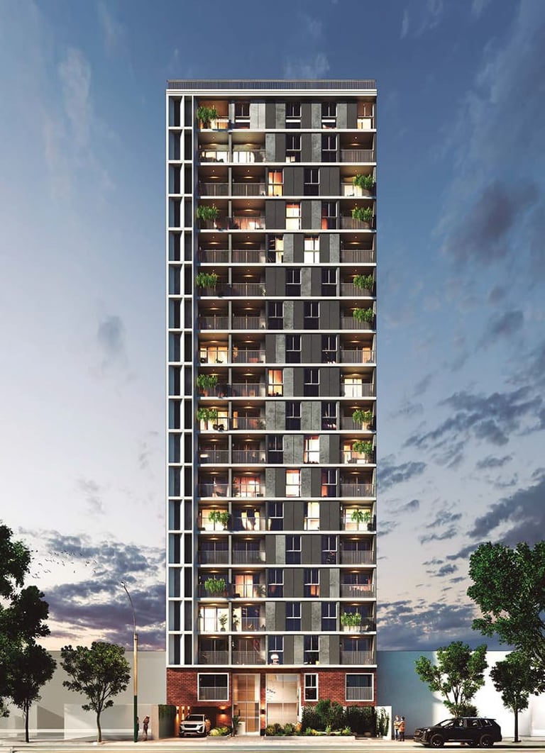 Modern residential high-rise apartment building with illuminated units, white steel frame, and landscaping at street level against cloudy sky.