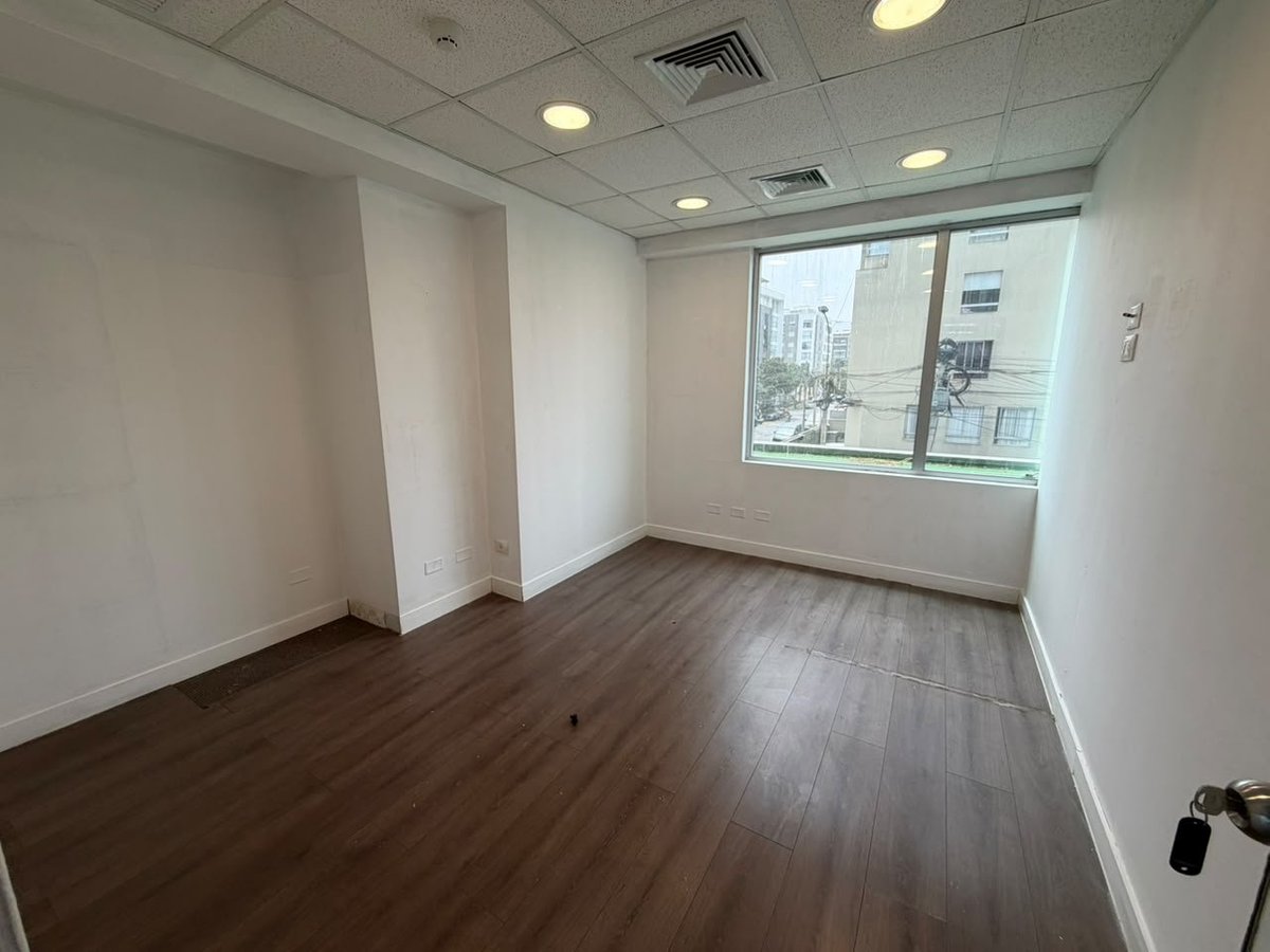 Empty modern office or apartment room with dark wood flooring, white walls, recessed ceiling lights, and large window overlooking city buildings