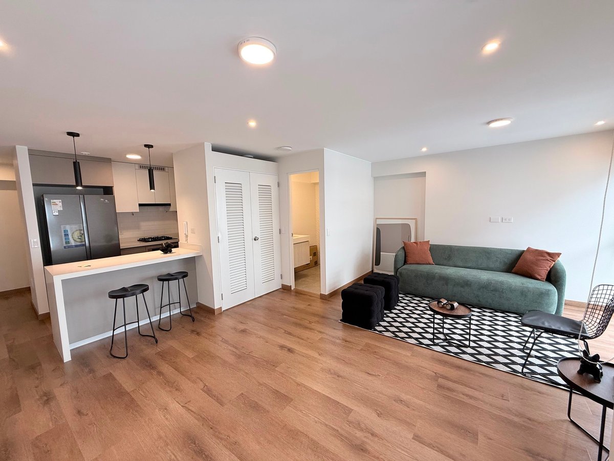 Open concept apartment with kitchen bar, seating area with green sofa, hardwood flooring, and modern white walls and fixtures