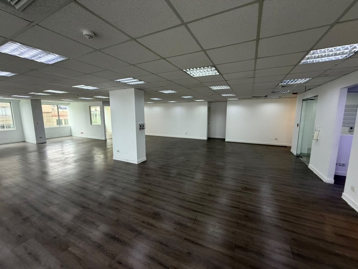 Empty modern office or retail space with dark wood flooring, white walls, support columns, fluorescent ceiling lights, and glass partitions.