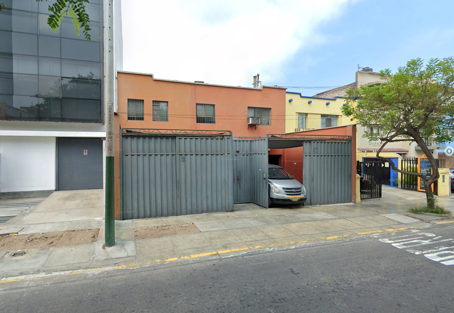 Urban street with parked car behind corrugated metal gates, orange and yellow residential buildings, and a modern glass structure on the left