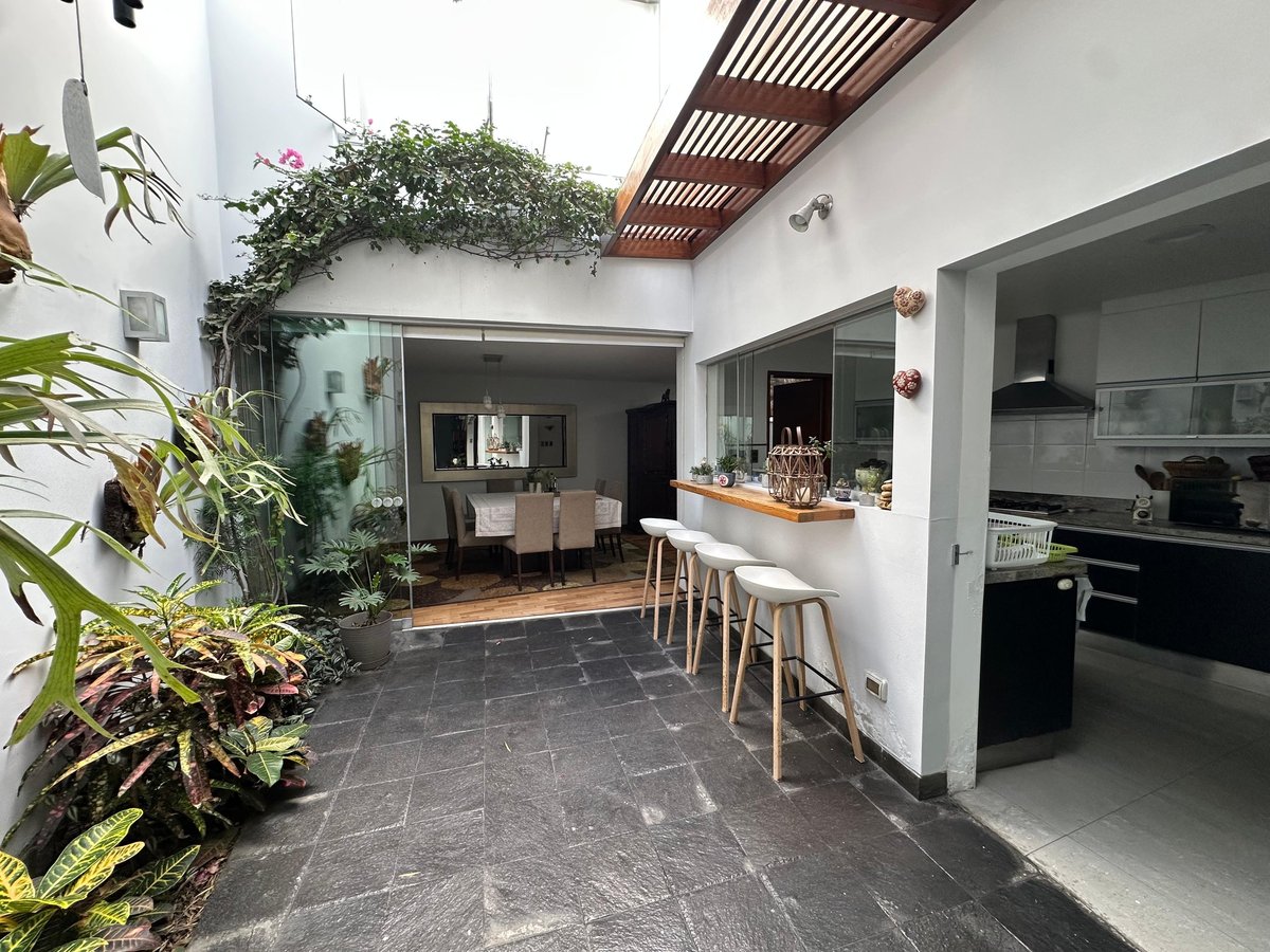Modern kitchen and dining area with dark flooring, white walls, wooden bar stools, and indoor plants in an open-concept home layout