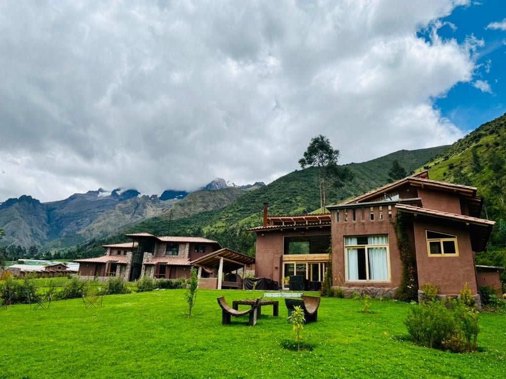 Rural lodge complex with terracotta buildings set on green grass beneath mountains and cloudy sky