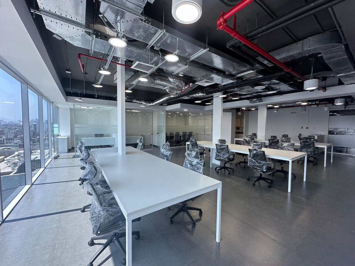 Modern open office space with white desks, office chairs, large windows, exposed ceiling with red piping, and city views