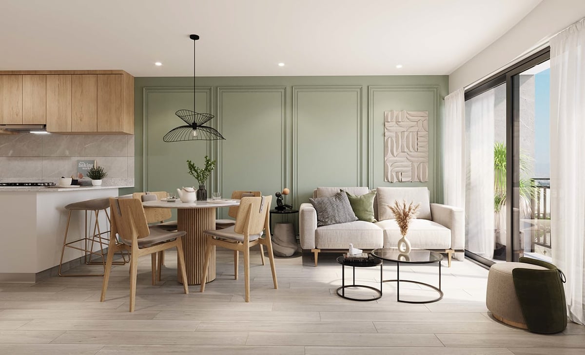 Modern open-concept apartment interior with light green walls, wooden dining table, beige sofa, kitchen with natural wood cabinets, and glass doors leading to a balcony