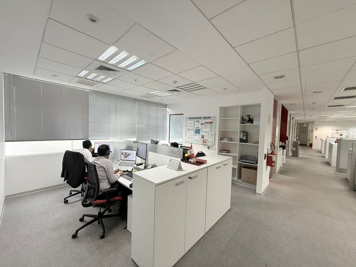 Modern office space with white cubicles, two employees working at desks, fluorescent ceiling lights, and shelving units along the walls