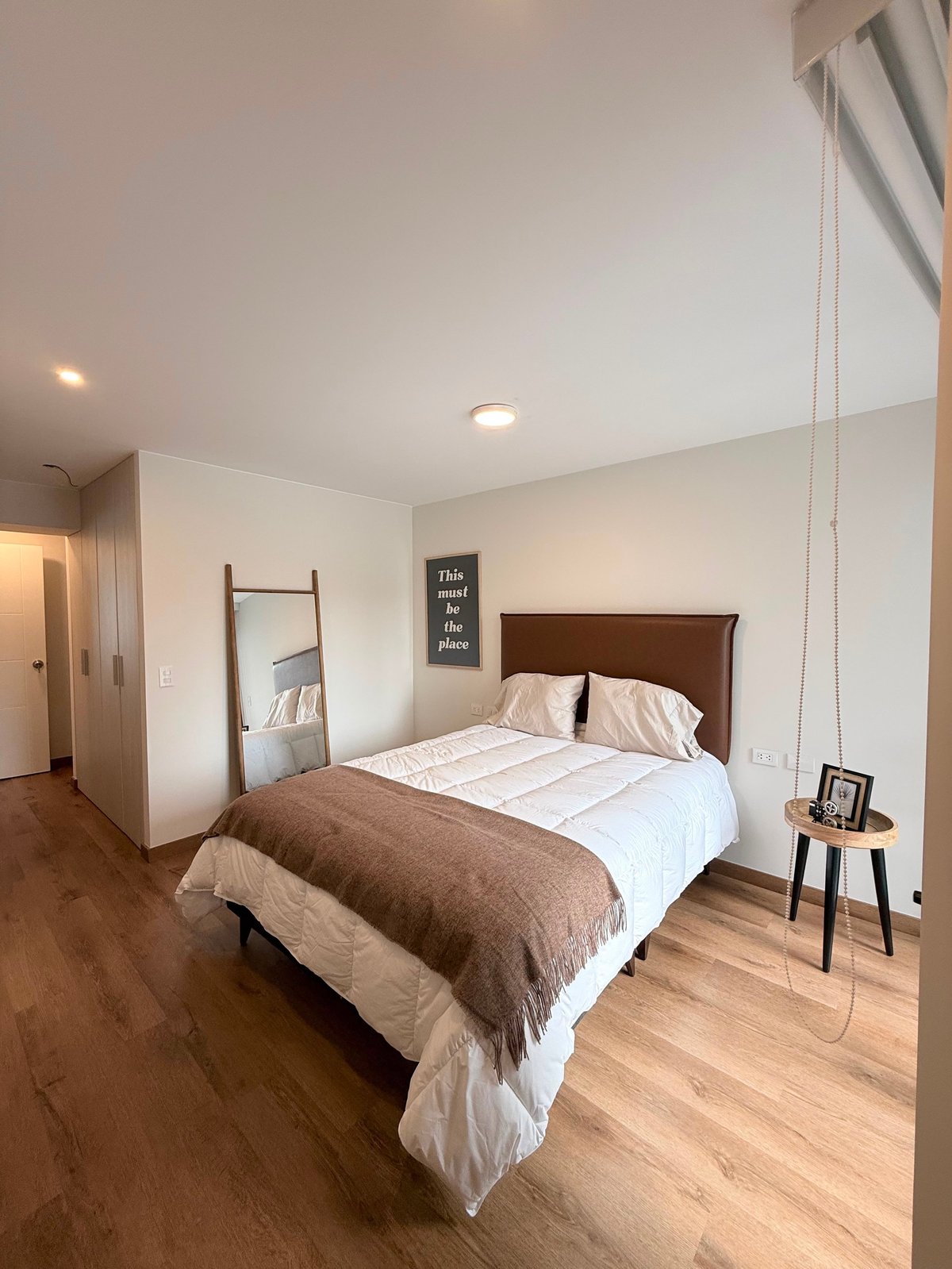 Modern bedroom with wooden bed frame, white bedding, brown throw blanket, wooden floors, and minimalist decor with wall signage