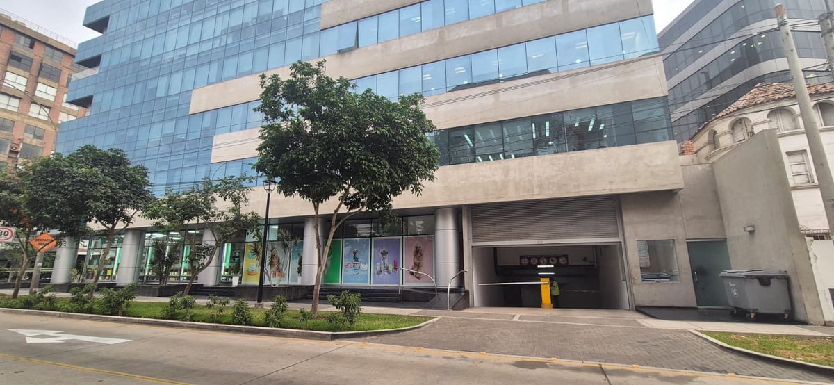 Edificio de oficinas moderno con fachada de vidrio y piedra, comercios a nivel de calle, área ajardinada y acceso a estacionamiento subterráneo
