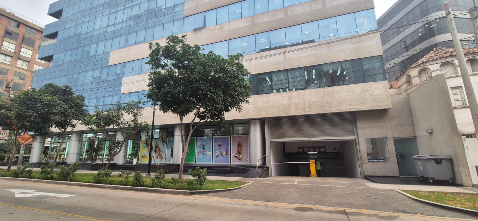 Edificio de oficinas moderno con fachada de vidrio y piedra, comercios a nivel de calle, área ajardinada y acceso a estacionamiento subterráneo