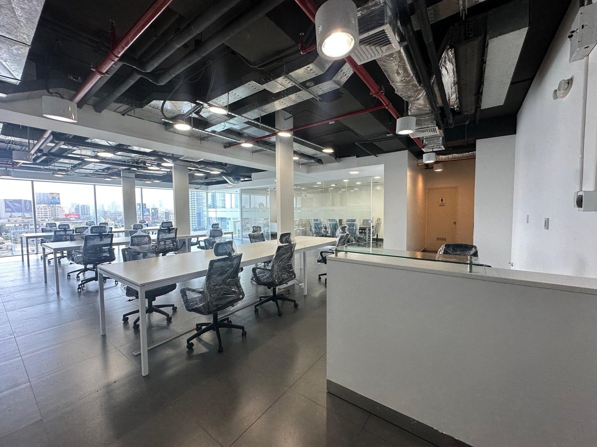 Modern open-plan office space with desks, office chairs, exposed ceiling infrastructure, and large windows overlooking a city skyline