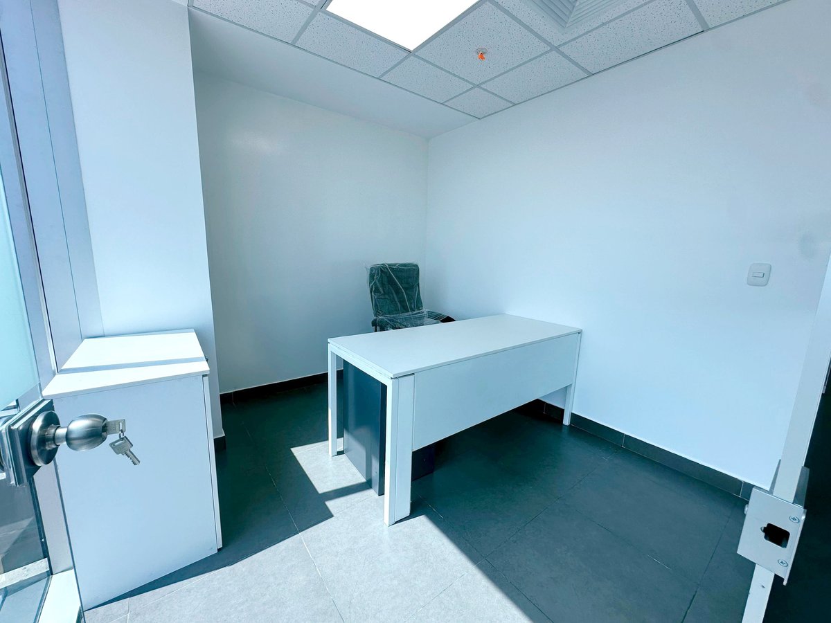 Empty minimalist office room with white desk, tiled ceiling, dark floor, and window on left wall