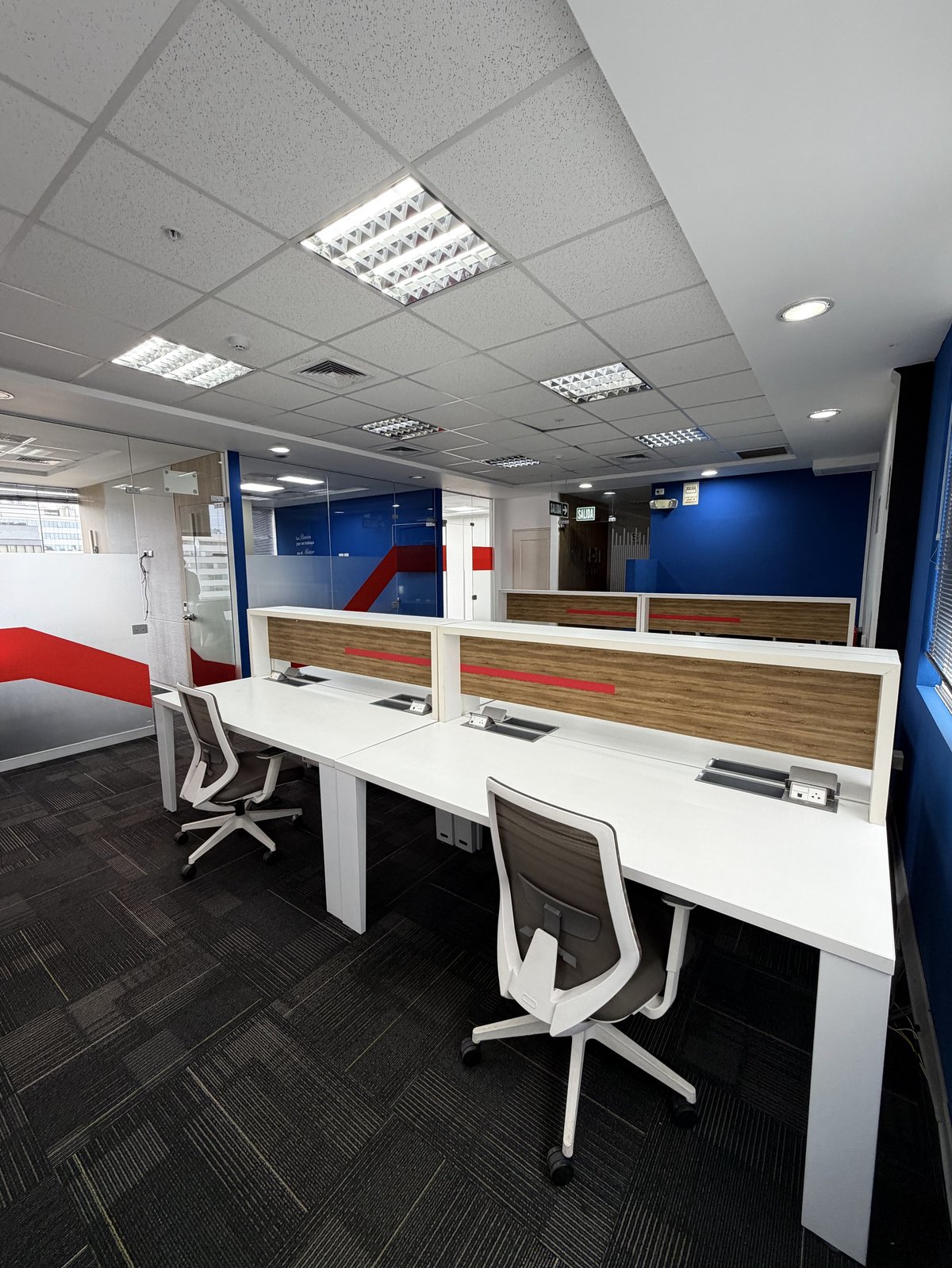 Modern office workspace with white desks, white ergonomic chairs, blue glass partitions, and fluorescent ceiling lights